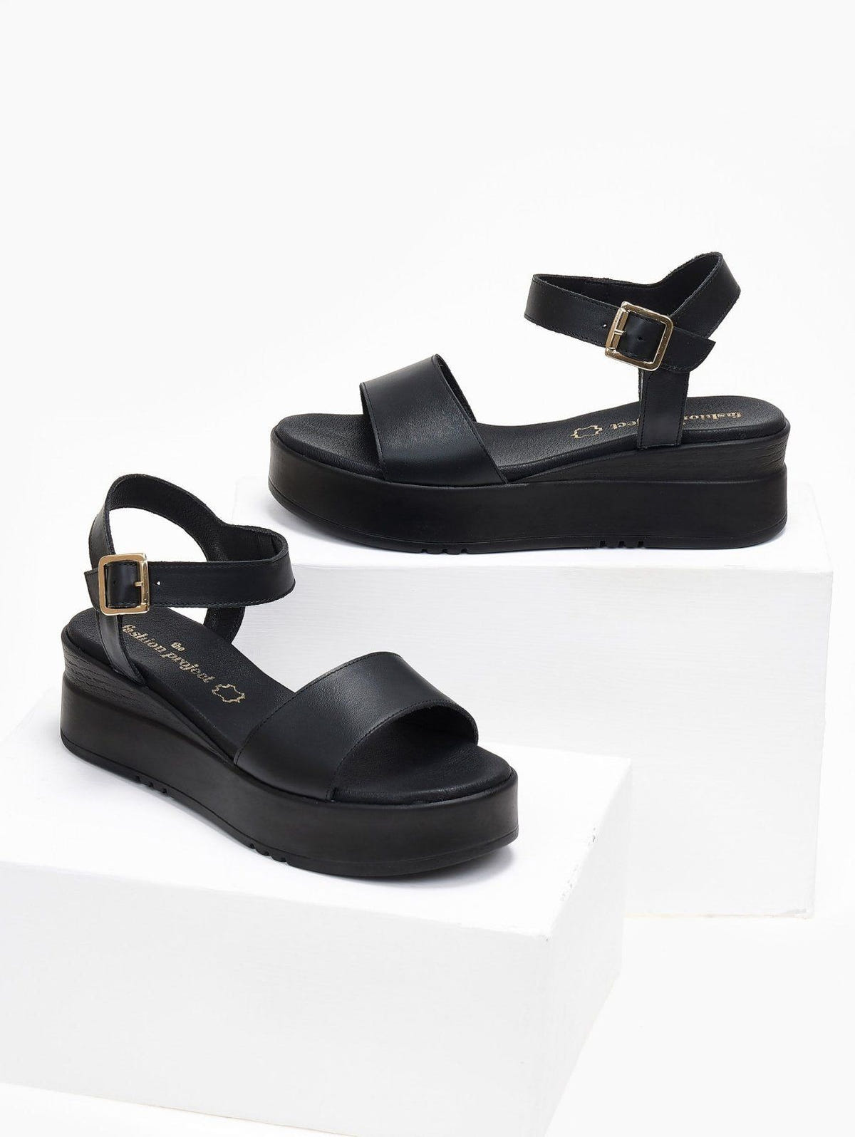 Pair of black leather flatform sandals with wide strap and ankle buckle, displayed on white blocks