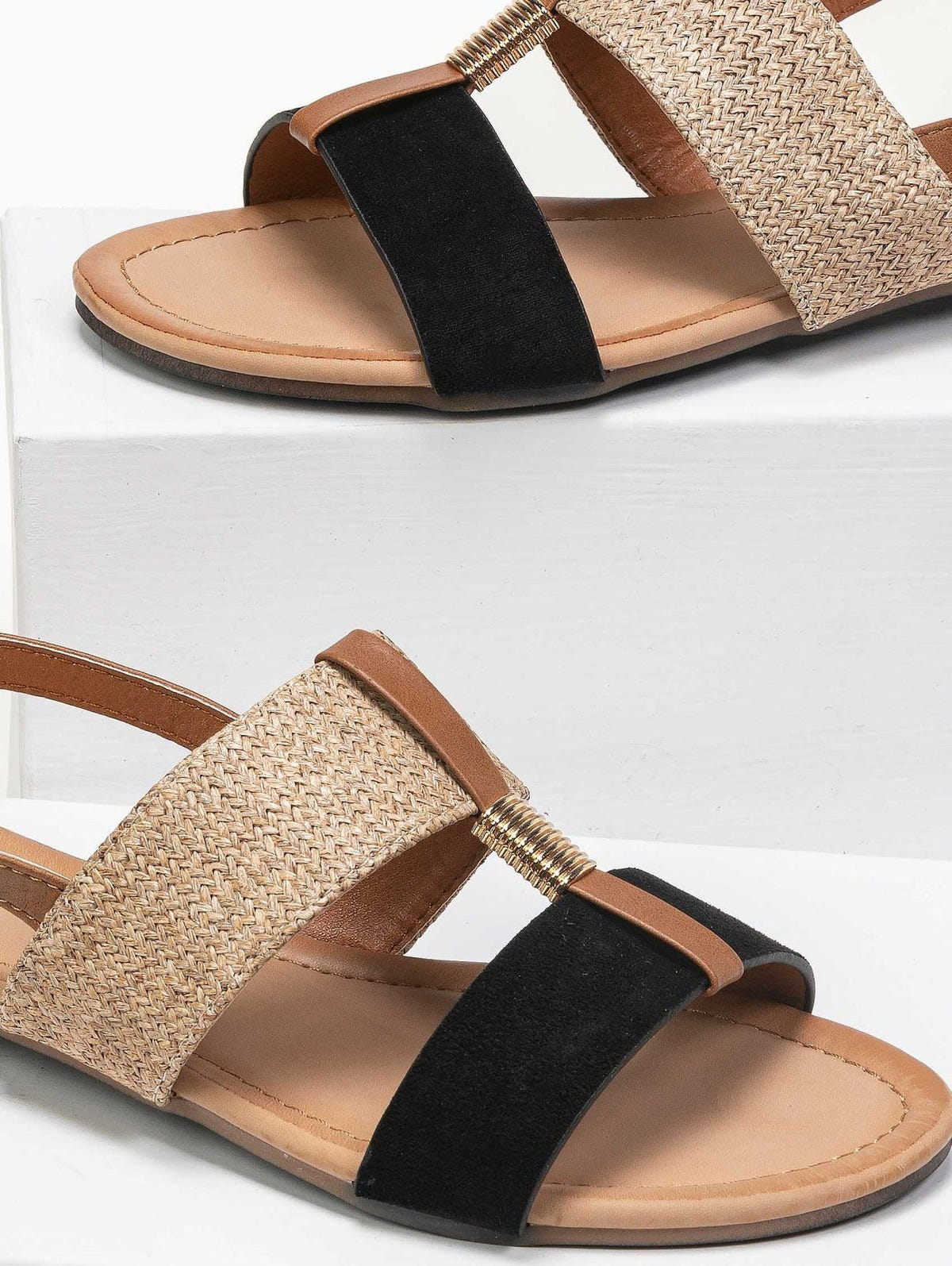 Close-up of black suede and woven straw flat sandals with tan leather trim, gold coil detail and tan insole, shown on white blocks
