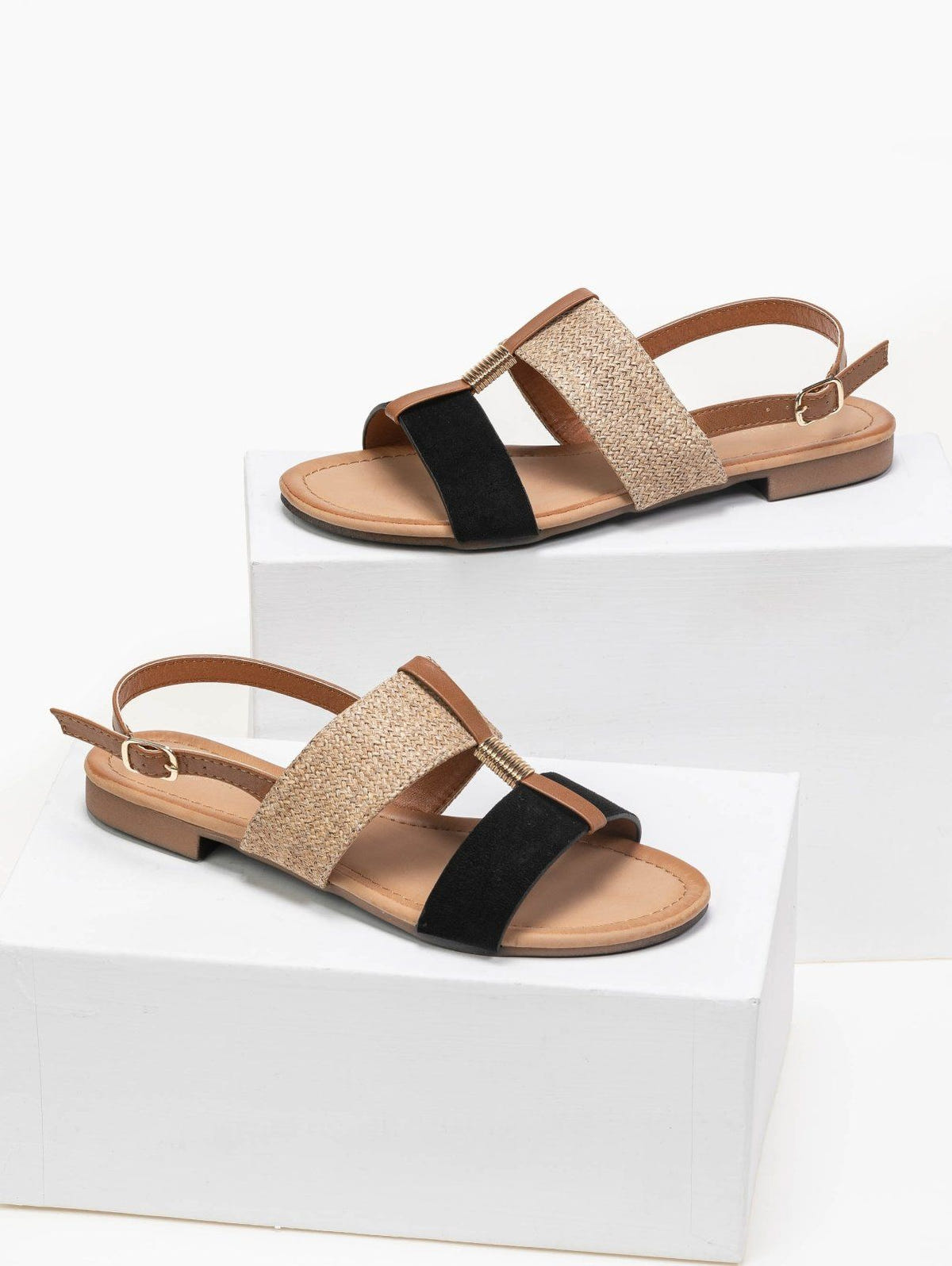 Pair of flat sandals with woven straw and black suede straps, tan leather slingback ankle strap with gold buckle, displayed on white blocks
