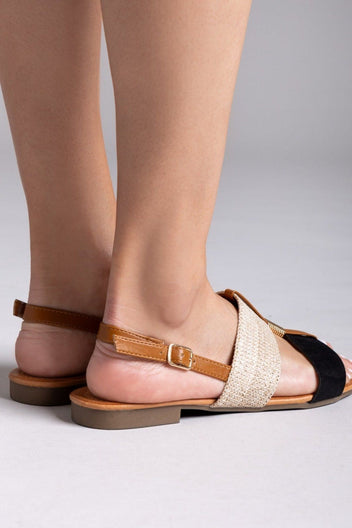 Back view of flat sandal on model showing tan leather ankle strap with gold buckle, woven straw band, black suede strap and low heel