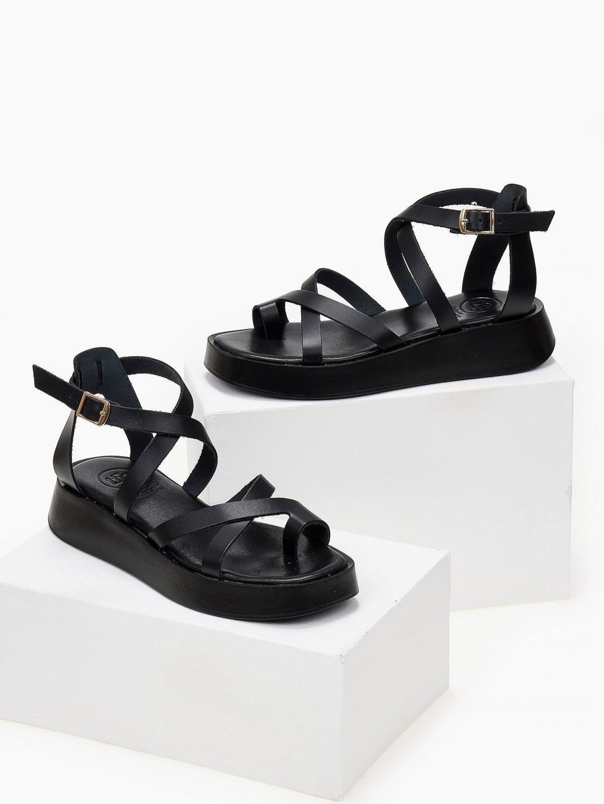 Pair of black leather crossover strap platform sandals with toe loop and ankle buckle, on white blocks