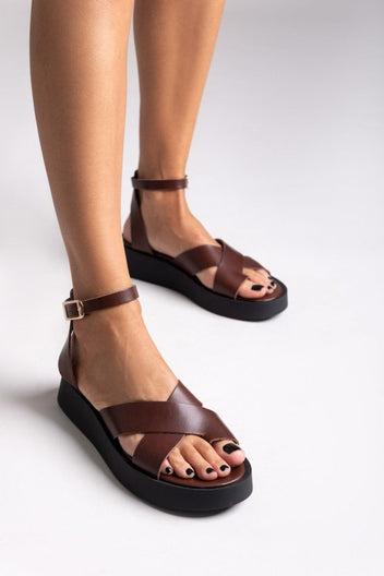 Model wearing brown leather crossover strap platform sandals with ankle buckle, front three-quarter view