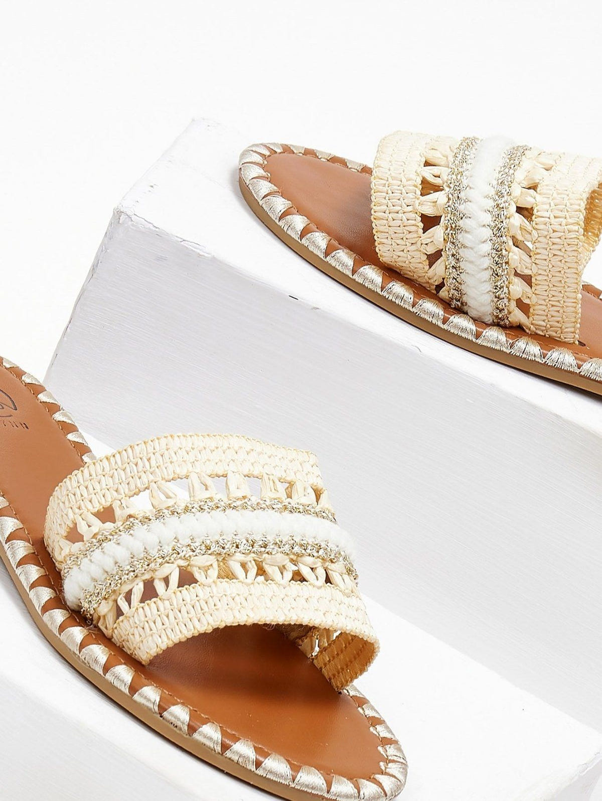 Close-up of natural raffia slide sandal with woven cream and white band on a white block