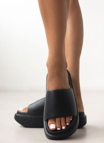 Model wearing black matte platform slide sandals with wide padded strap and thick sole, front view