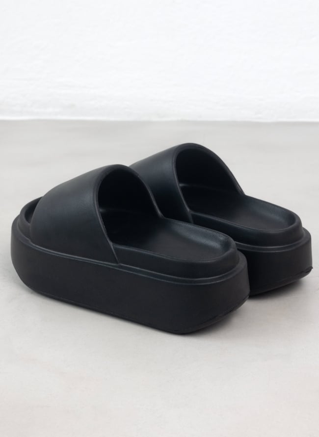 Pair of black chunky slides with wide padded straps and thick sole seen from the rear three quarter view