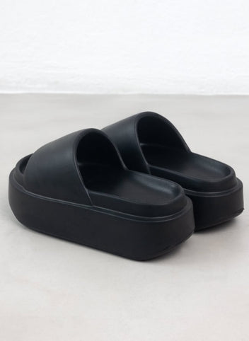 Pair of black chunky slides with wide padded straps and thick sole seen from the rear three quarter view