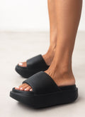 Model feet wearing black chunky slides with a wide strap and thick sole seen from the side