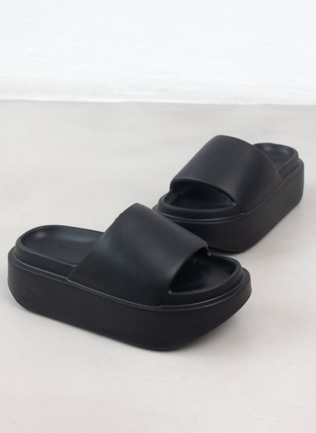 Front three quarter view of a pair of black platform slides with a wide strap and thick rubber sole on a pale surface