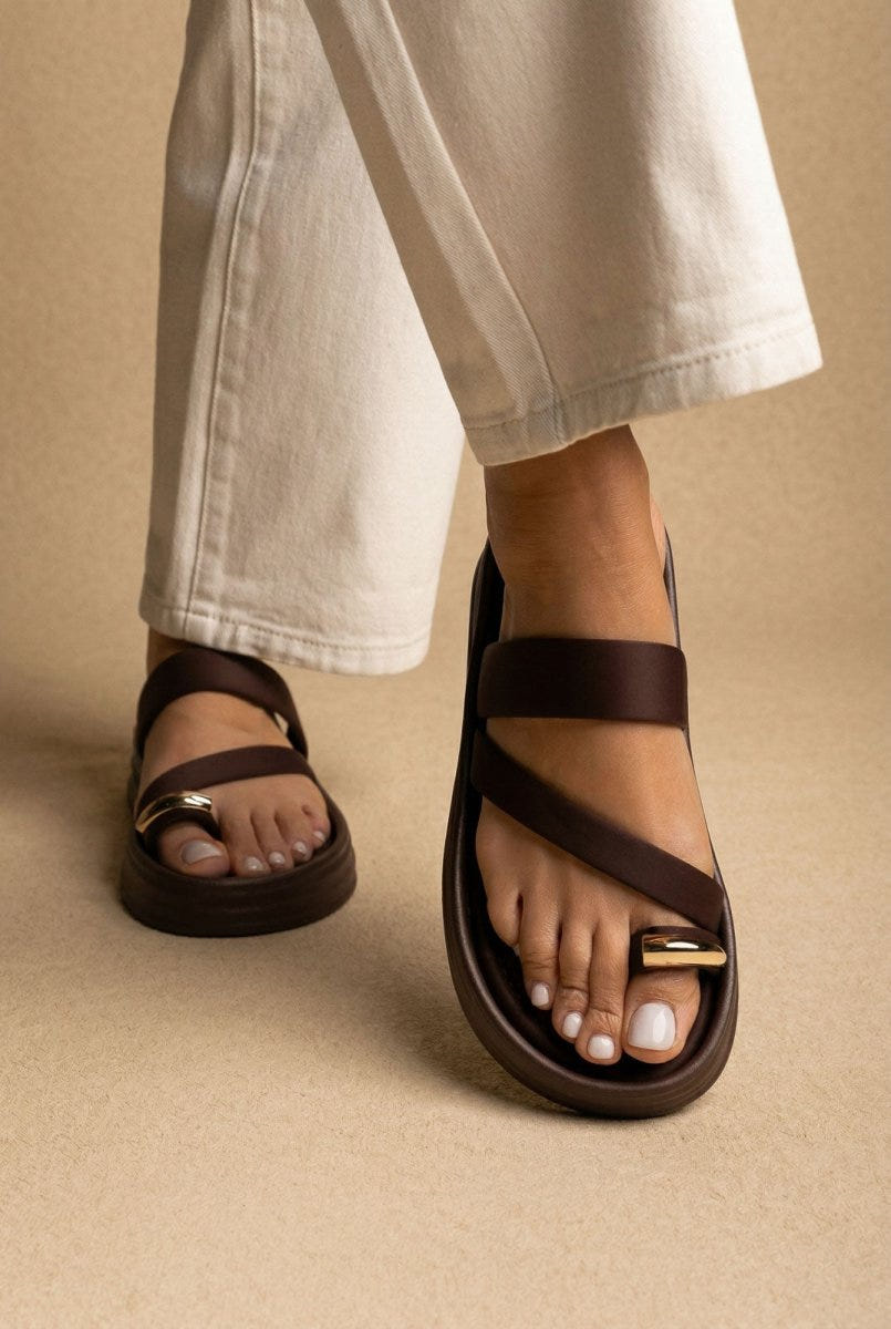 Model wearing brown flatform sandals with a wide strap and toe ring, close-up of the foot below white wide leg trousers