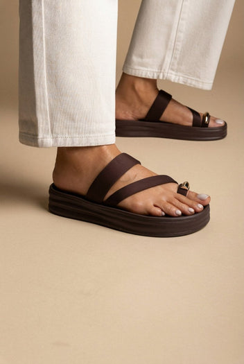 Model wearing brown satin flatform toe ring sandals with a metal ring, legs in cream jeans on a sand-toned surface