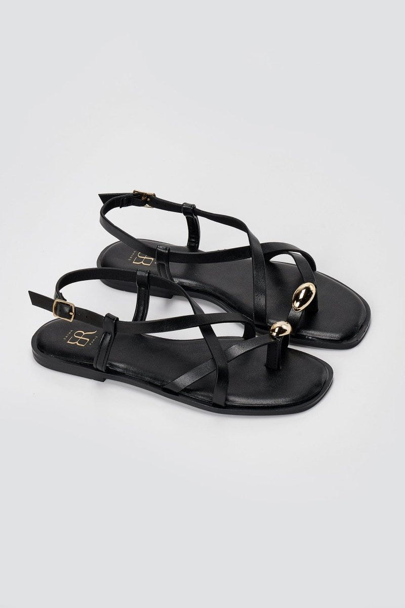 Pair of black leather toe ring sandals on light grey, three-quarter view showing ankle strap and gold ring at toe