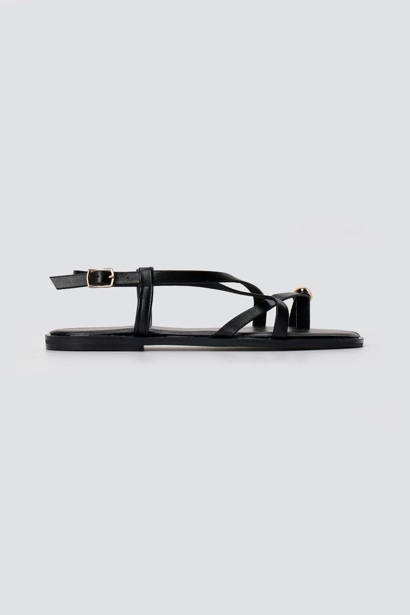 Black leather toe ring sandal in side view with thin straps, ankle buckle and a flat sole on light grey