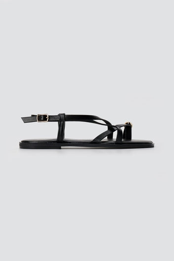 Black leather toe ring sandal in side view with thin straps, ankle buckle and a flat sole on light grey