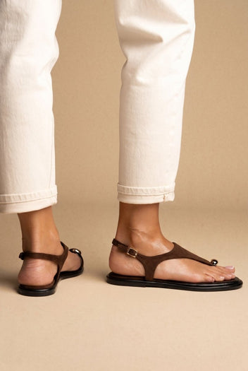 Model wearing brown suede thong sandals with black soles, lower legs in cream trousers and a metal ring at the toe