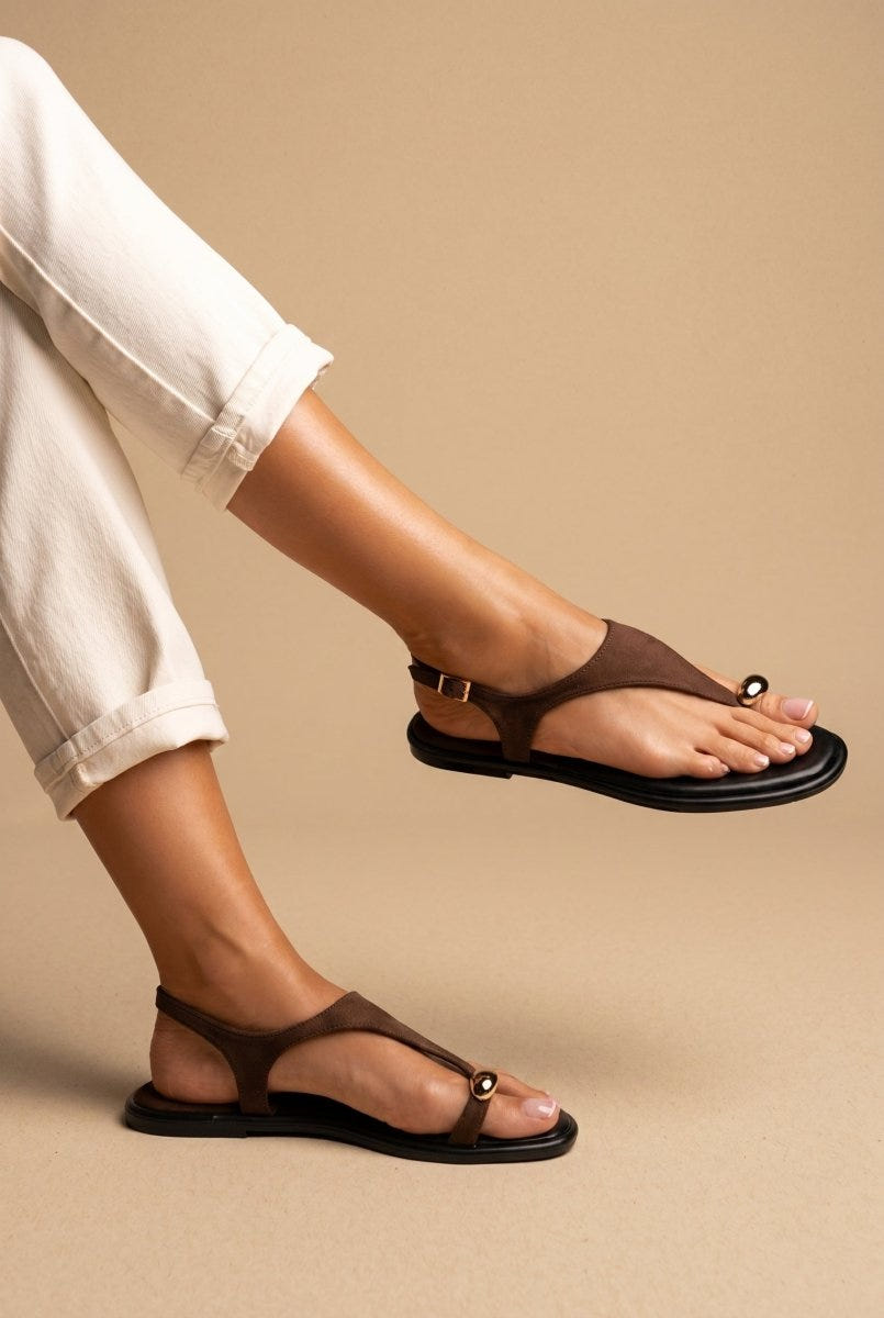 Model wearing brown suede thong sandals with a slim ankle strap, close-up of the foot in cream trousers on sand tones
