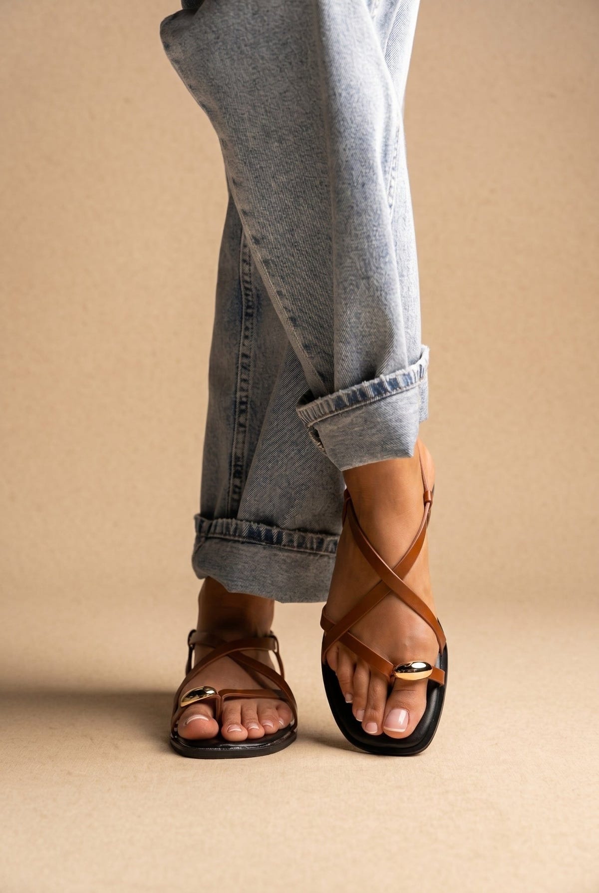 Model wearing tan leather toe ring sandals with cuffed grey jeans, low view of feet with straps and gold ring at toe